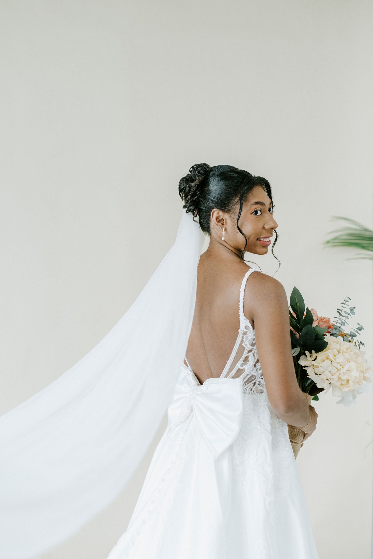 A bride in a flowing white wedding dress and veil holds a bouquet while looking over her shoulder.
