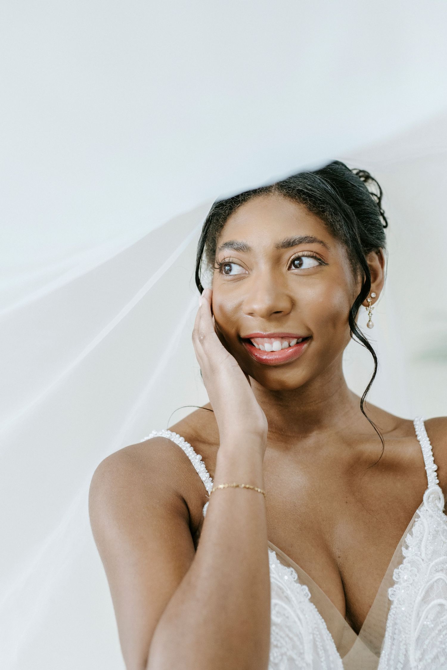 A bride in a white lace dress smiles joyfully while touching her face during a bridal portrait session.