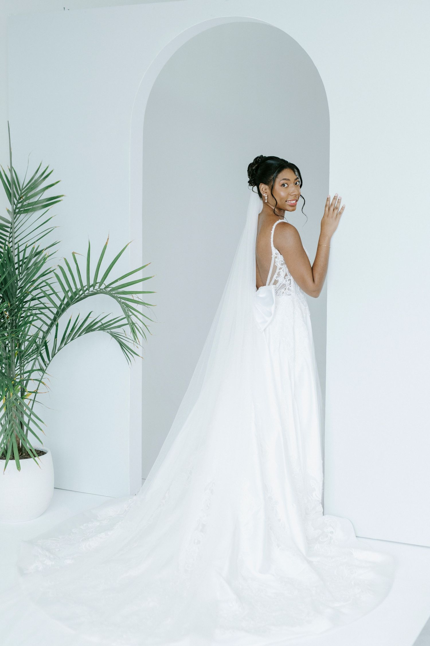 A minimalist bridal photoshoot featuring a flowing white wedding gown against a light background with palm fronds.