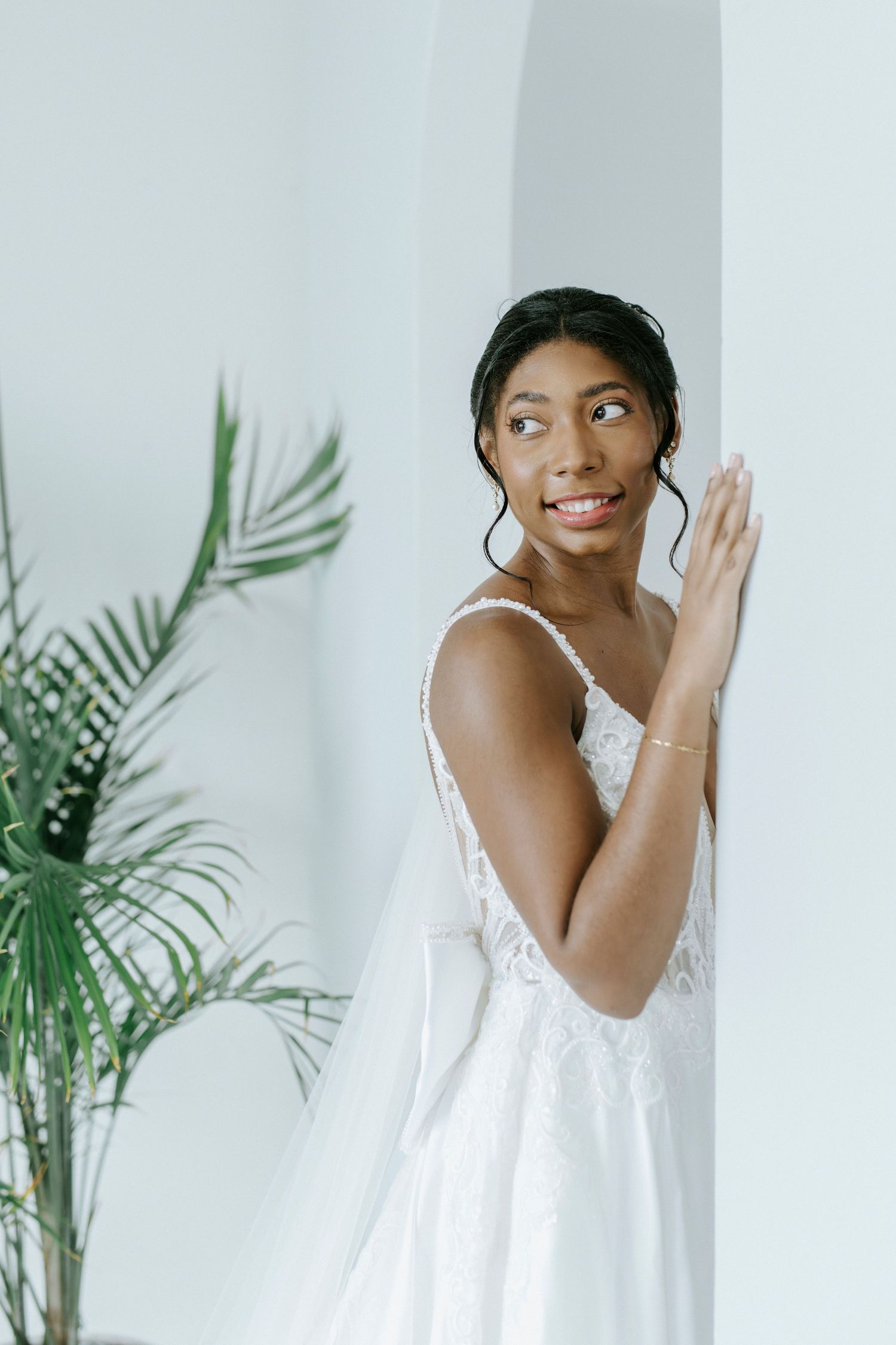 A bride in a delicate white gown poses gracefully against a light backdrop with tropical greenery.