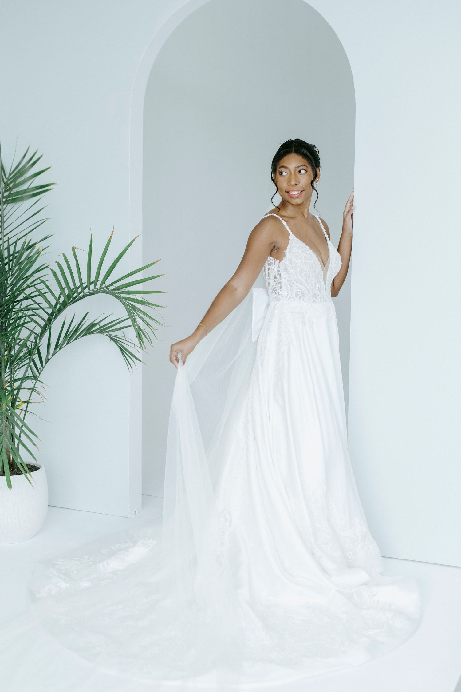 Ethereal bridal photo in flowing white gown with spaghetti straps against minimalist white archway and palm decor.