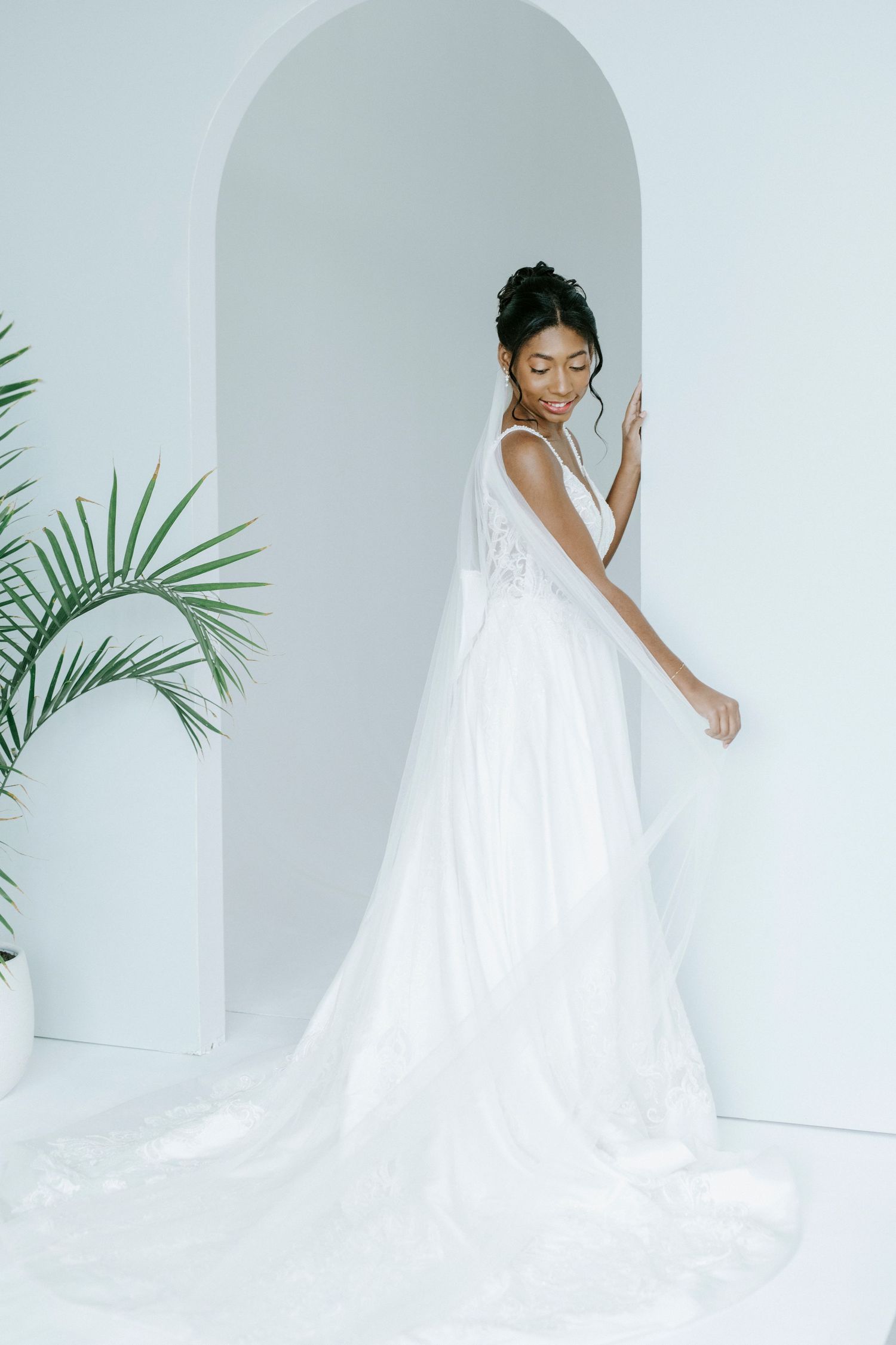 A bride in a flowing white dress twirls and dances in an elegant white studio setting with palm leaves.