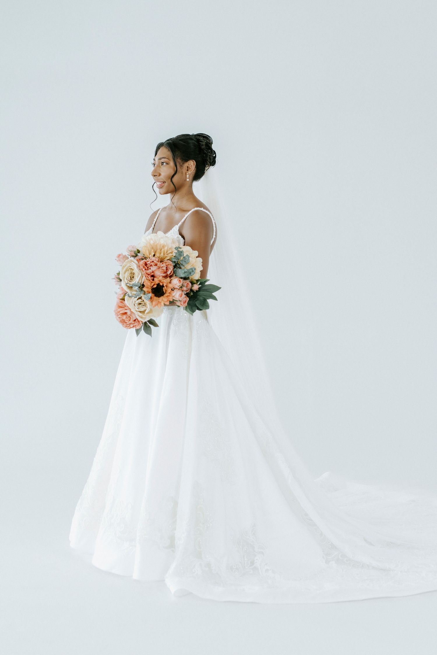 A bride in a flowing white dress holds a peach and coral bouquet against a minimalist light background.