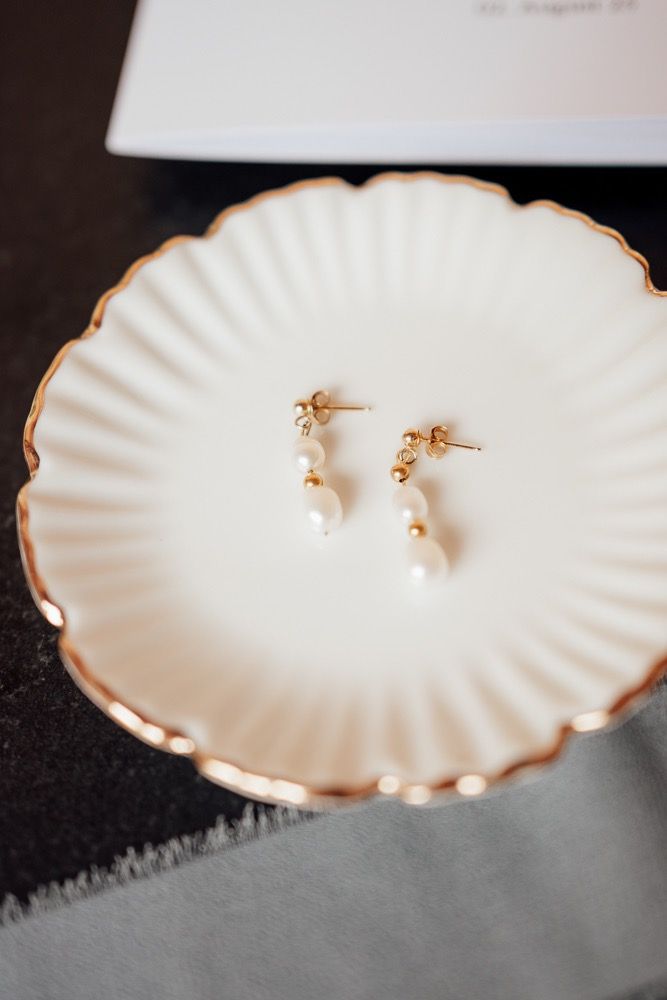Delicate pearl earrings displayed on white scalloped dishes with gold trim.
