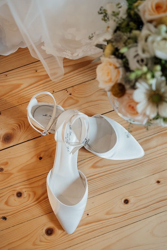 White bridal shoes with ankle straps displayed on a rustic wooden floor next to floral arrangements.