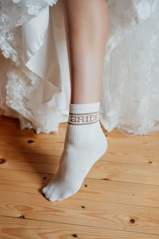 White lace wedding dress hem and pearl-adorned bridal sock on wooden floor.