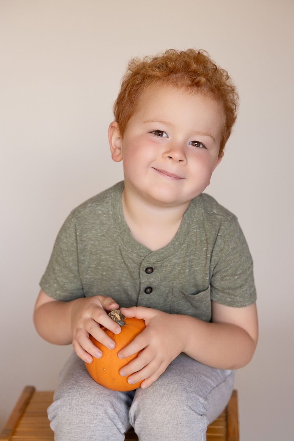 A young child holds a pumpkin while sitting on a stool for his preschool portraits