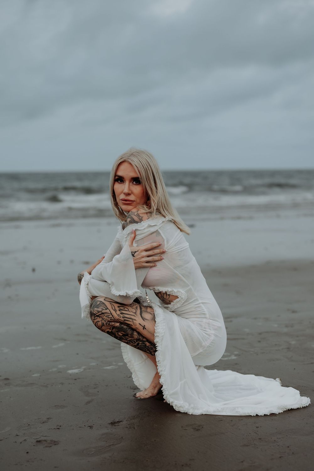 Person in flowing white dress crouches on sandy tannum sands beach during moody overcast weather.