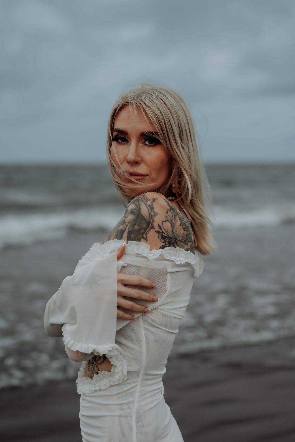 Woman in white dress poses dramatically on moody gladstone beach with stormy skies in background.