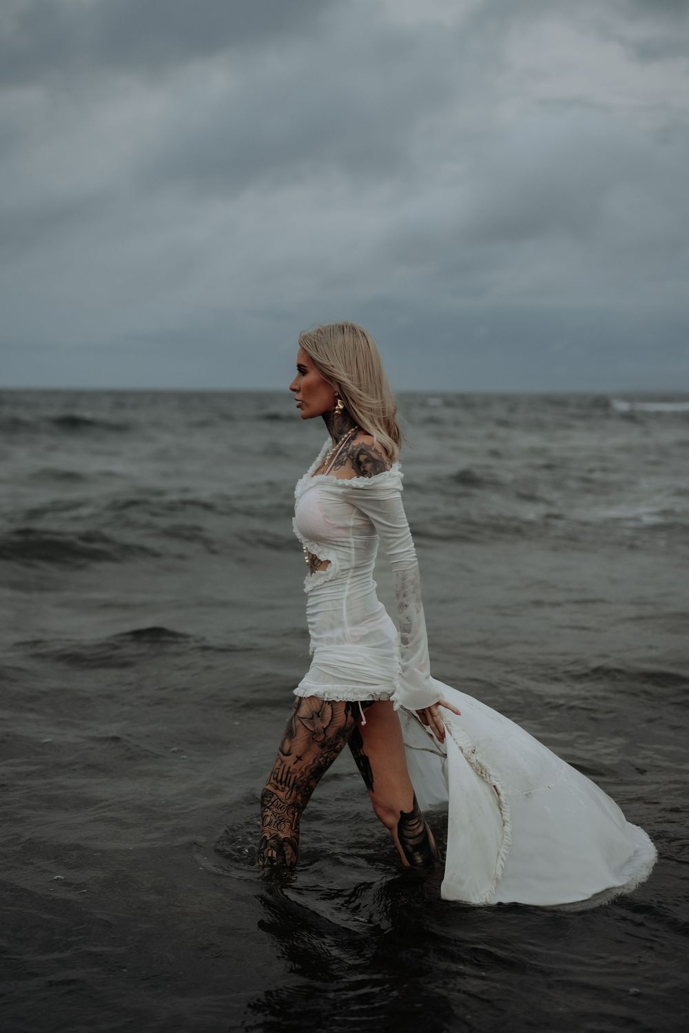 Figure in flowing white dress wades through dark ocean waters under cloudy sky at gladstone beach.