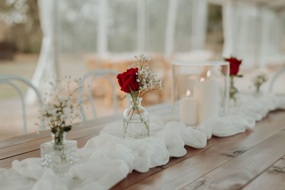 Red roses in small glass vases line a rustic wooden table with white fabric runner.