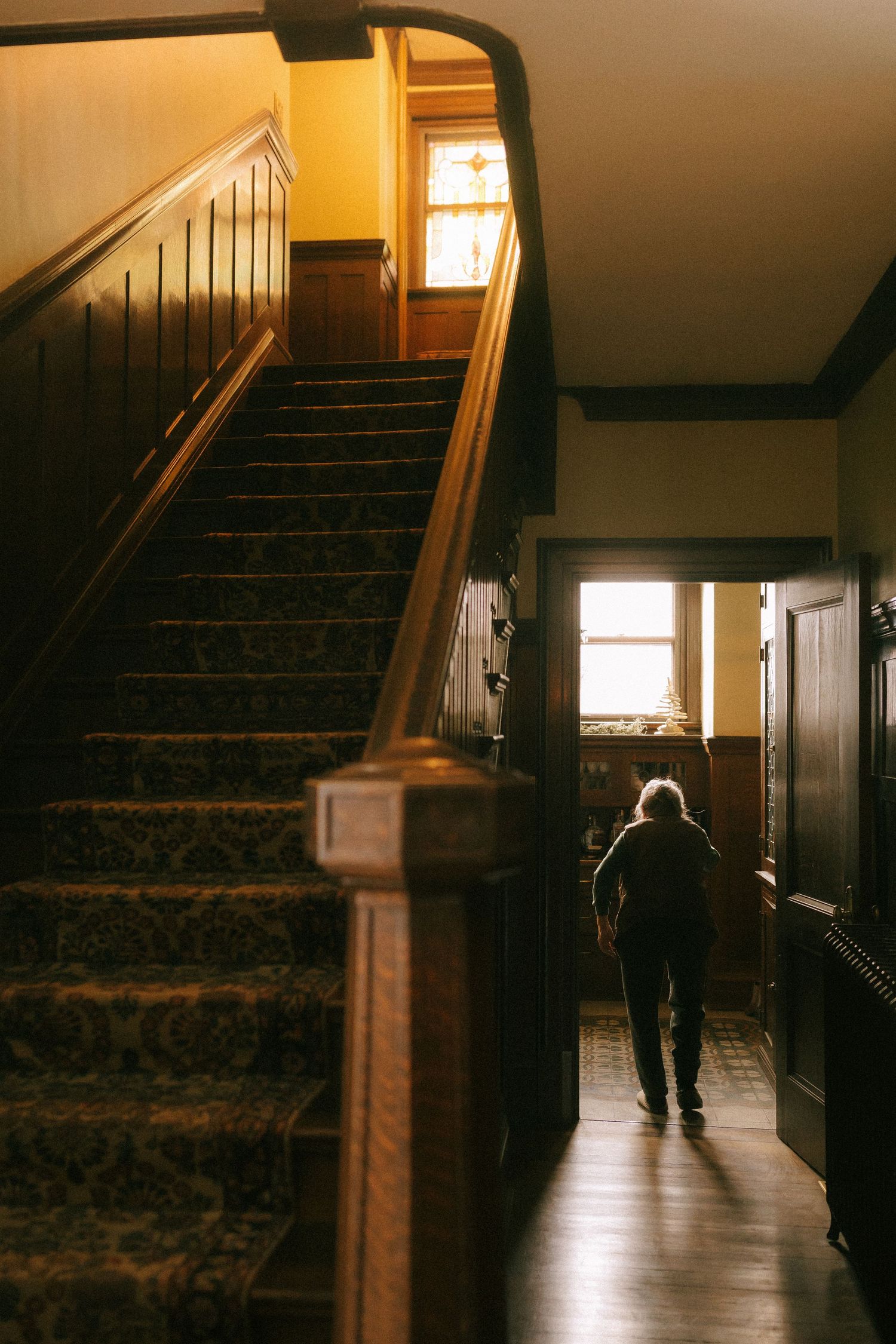 A silhouetted figure walks down a dimly lit hallway with wooden stairs and warm lighting from windows casting shadows.