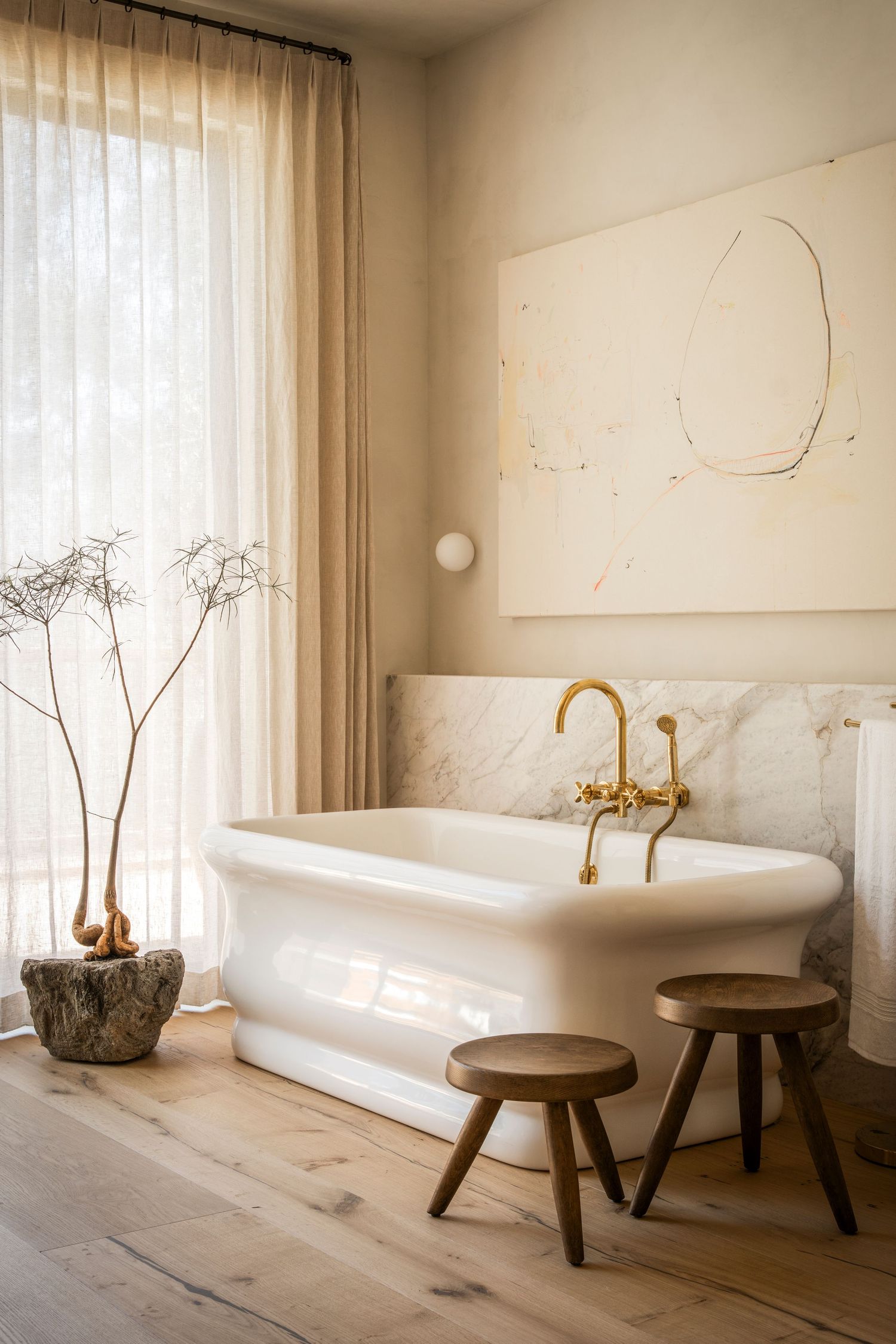 Minimalist bathroom with freestanding tub, brass fixtures, wooden stools, and sheer curtains in warm neutral tones.