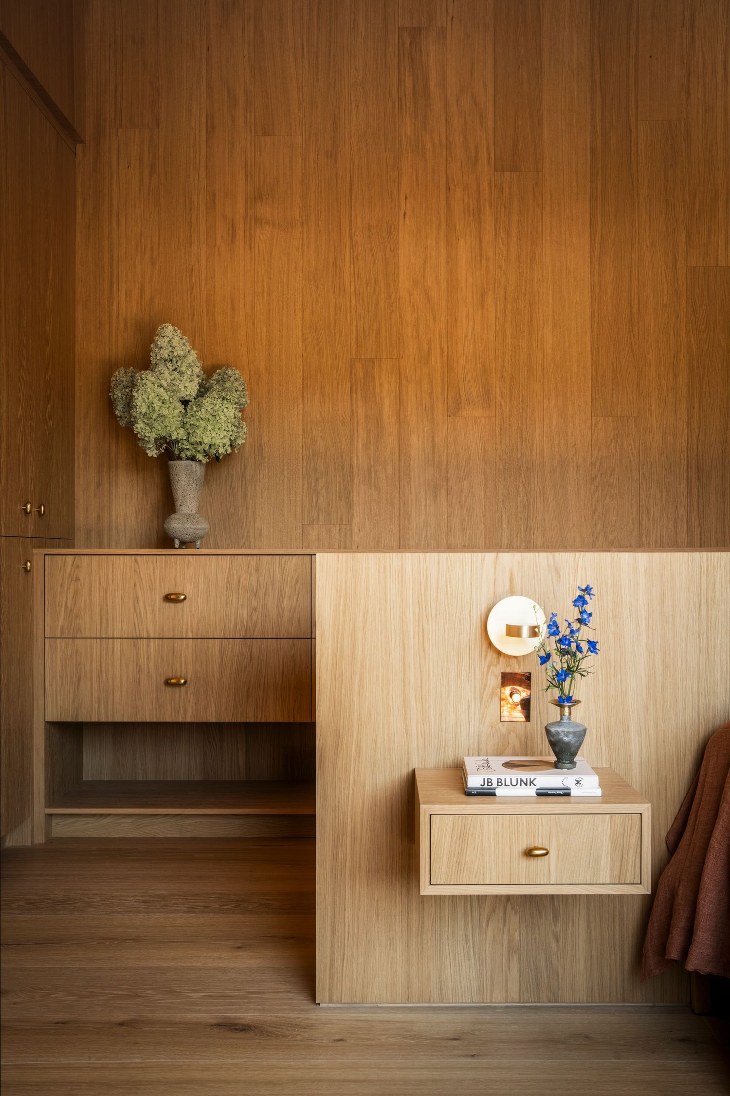 Modern wooden interior with built-in drawers and floating shelf decorated with hydrangeas and delphinium flowers in vases.