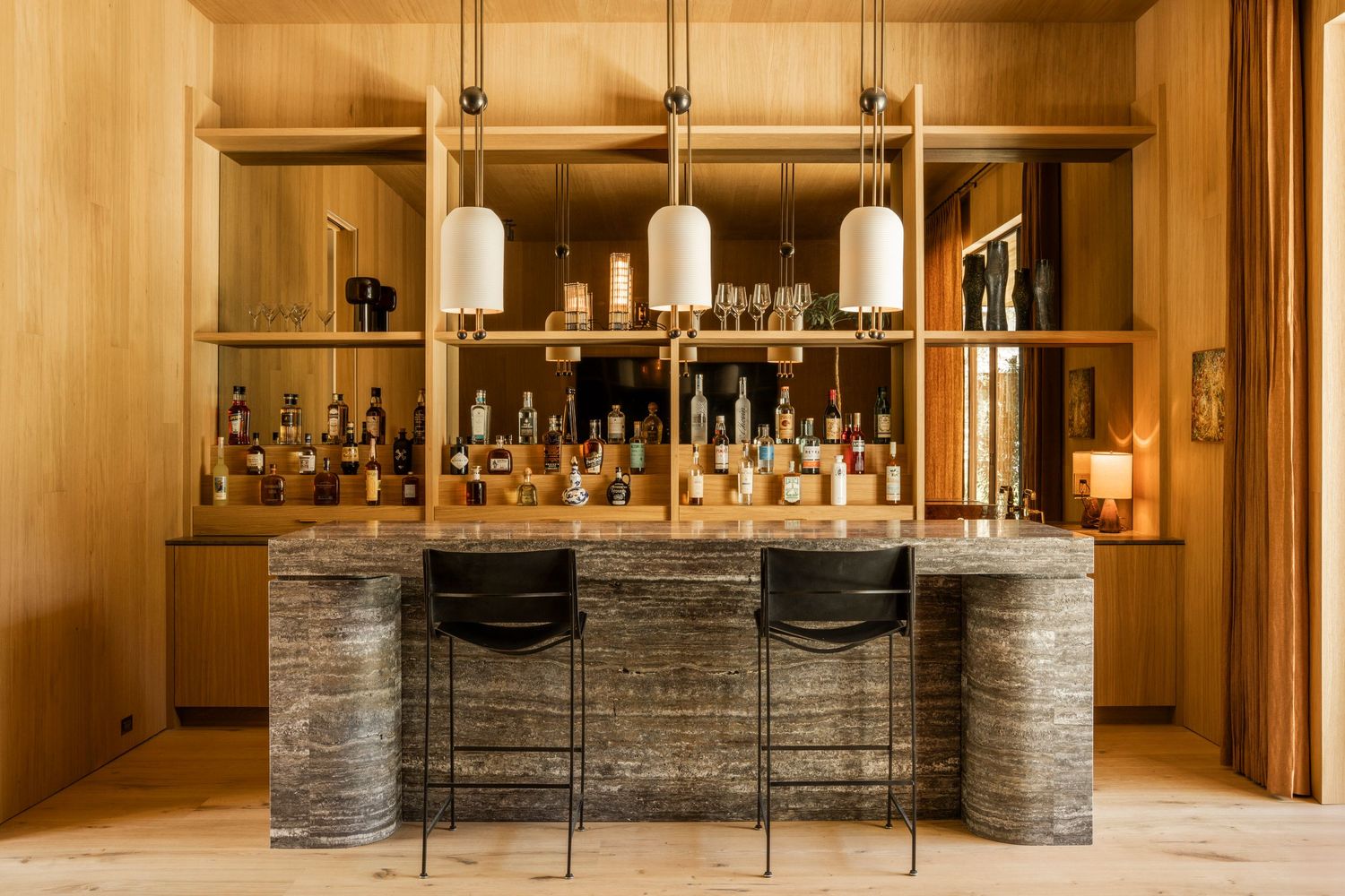 A modern hotel bar with stone pillars and warm lighting features sleek wooden surfaces and glass bottle displays.
