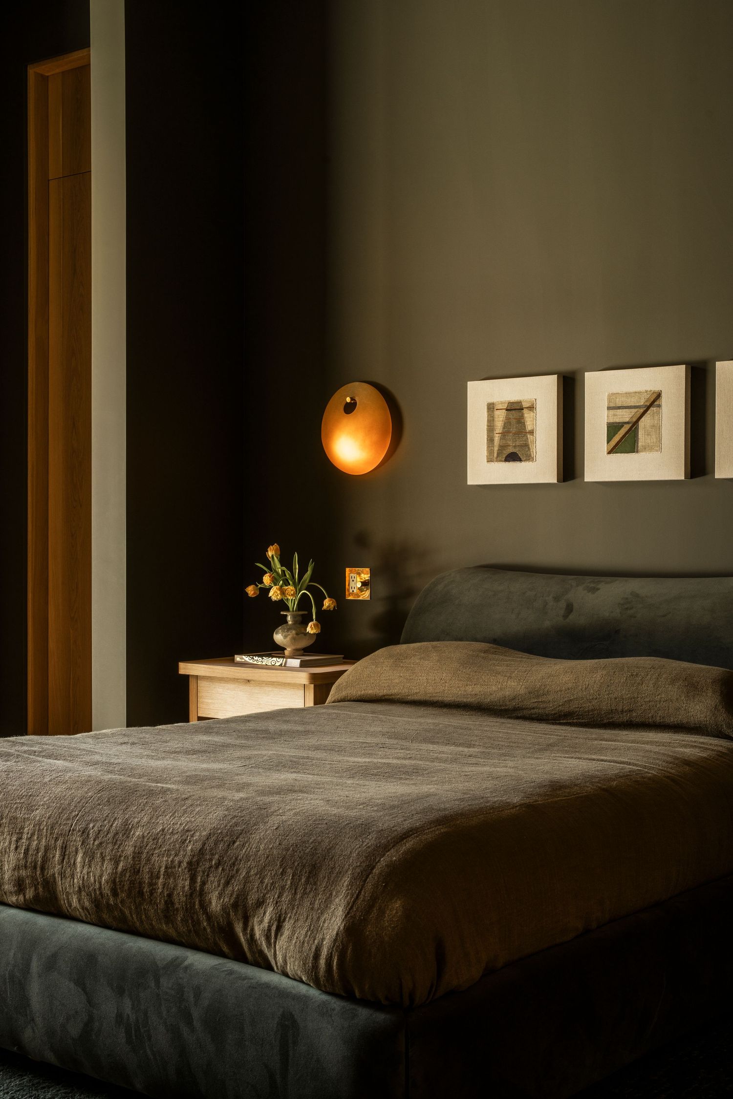 A cozy modern bedroom with dark walls features a bed with linen bedding, wall sconce lighting, and framed artwork.