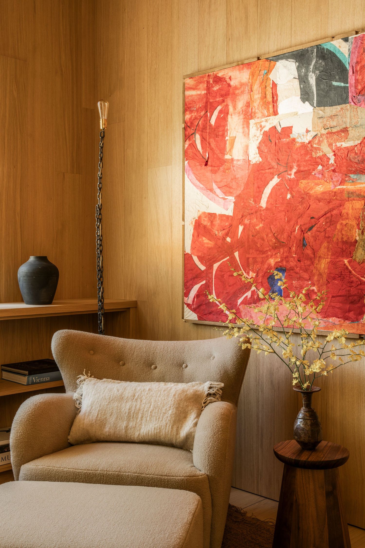 Modern living room corner with beige armchair, red abstract artwork, and wooden accent table featuring floral arrangement.