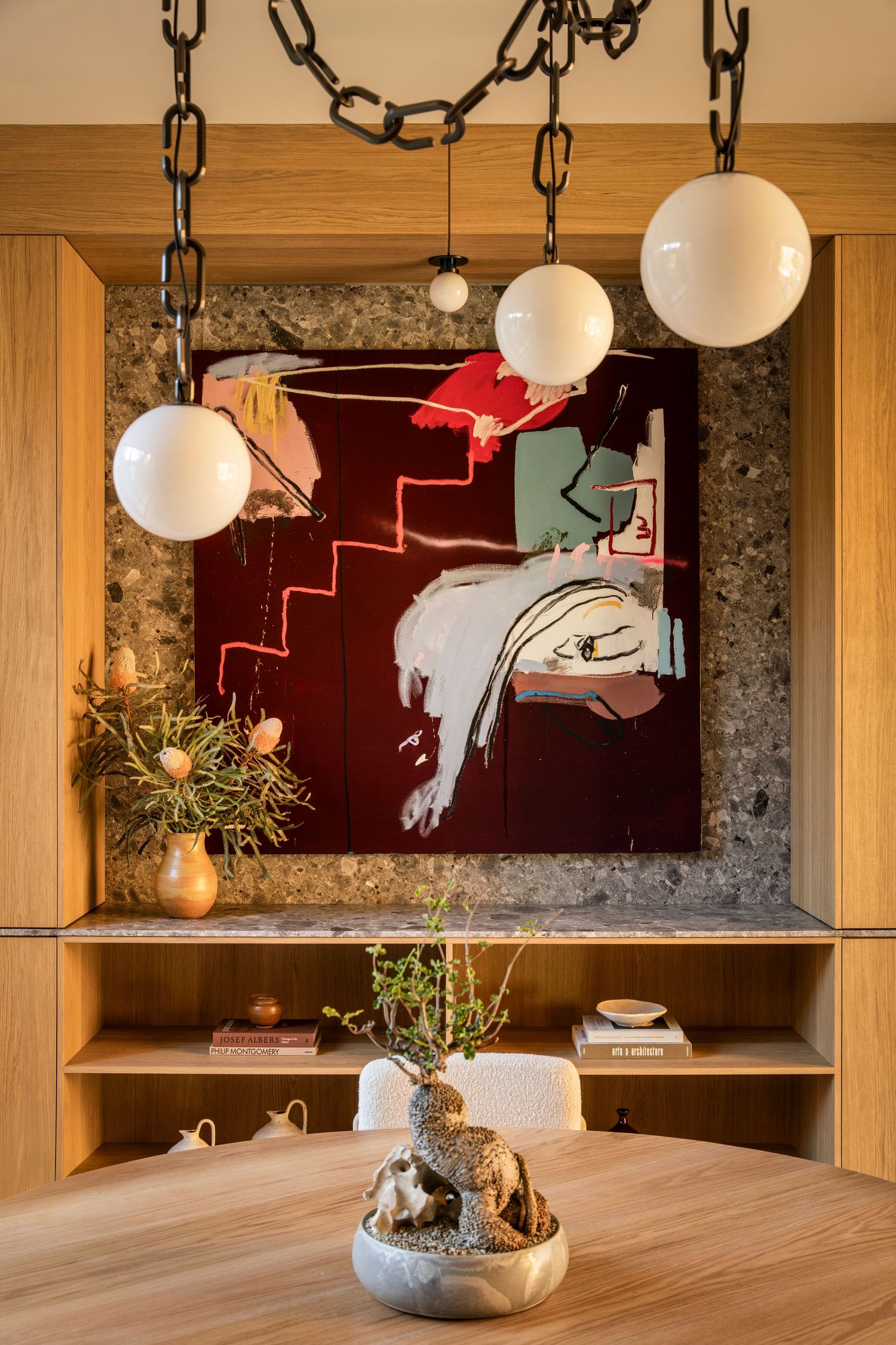 Modern dining room interior with wooden table, hanging pendant lights, and red abstract artwork on gray stone wall.