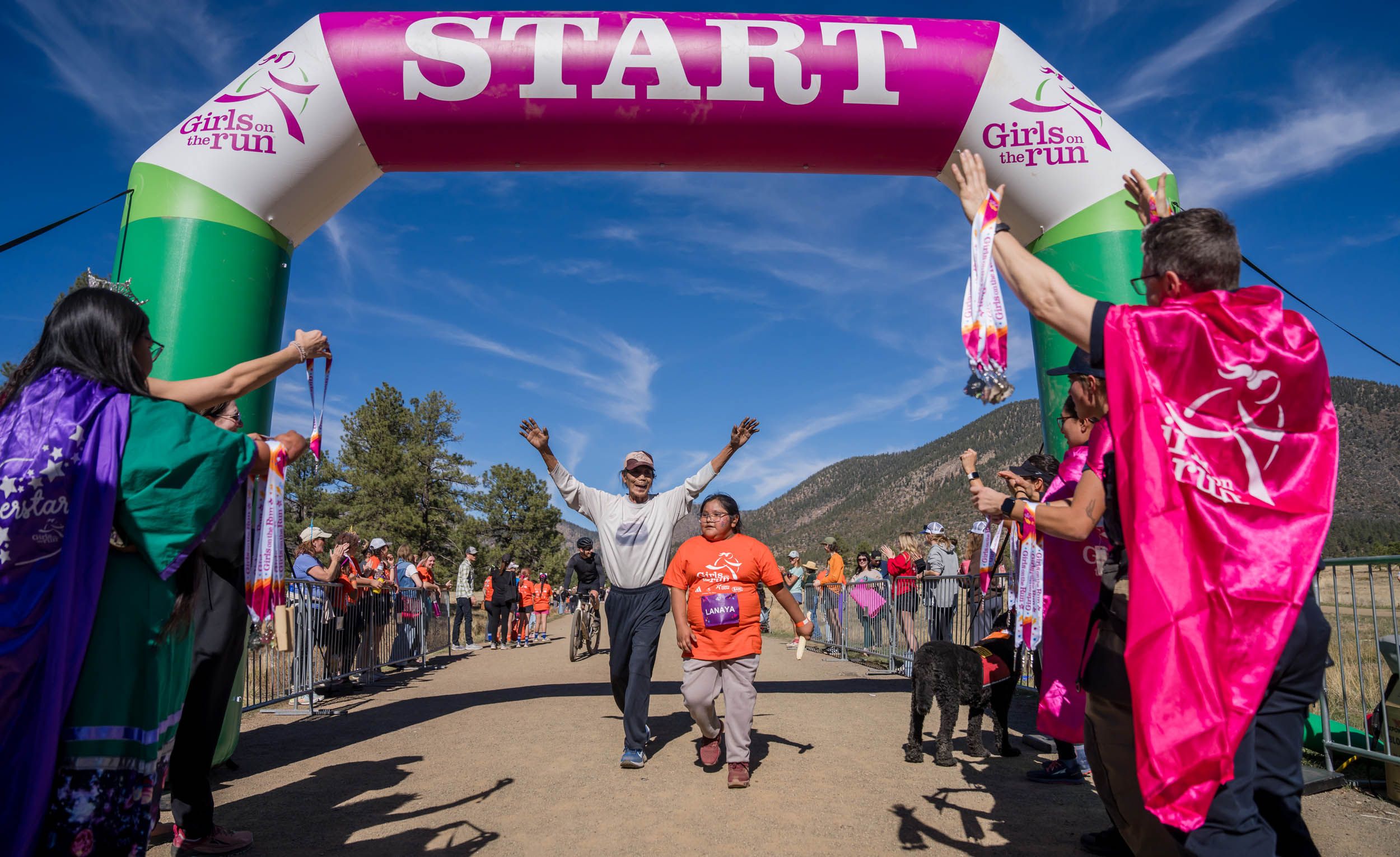 A series of pink and green flags wave along a desert trail with people gathering under bright blue skies.