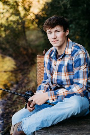 A person in a plaid shirt and jeans sits casually on a wooden surface during a golden hour outdoor photoshoot.