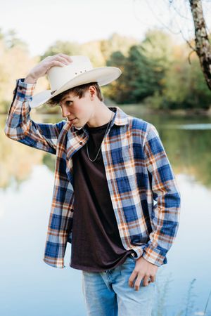 Person in plaid flannel shirt and cowboy hat poses by a serene lake during golden hour.