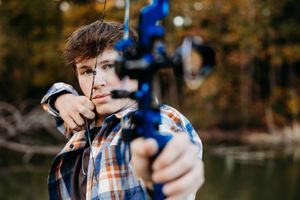 Person in blue and brown clothing aims a slingshot while standing in front of a blurred autumn forest background.