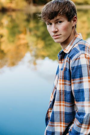 Young person in a plaid shirt and denim vest poses outdoors against a blurred autumn background with water reflections.