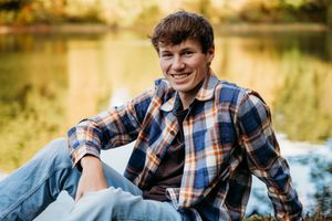 A person in a plaid shirt sits casually by a serene pond on an autumn day with golden light in the background.