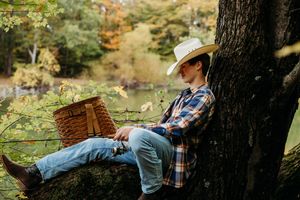 A person in a plaid shirt and cowboy hat rests peacefully against a large tree trunk in a serene forest setting.