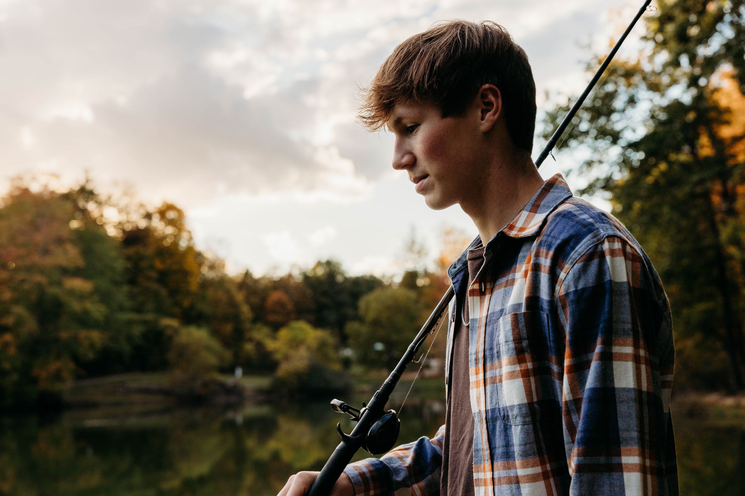 A person in a plaid shirt stands in a golden-lit field during sunset holding a fishing rod.