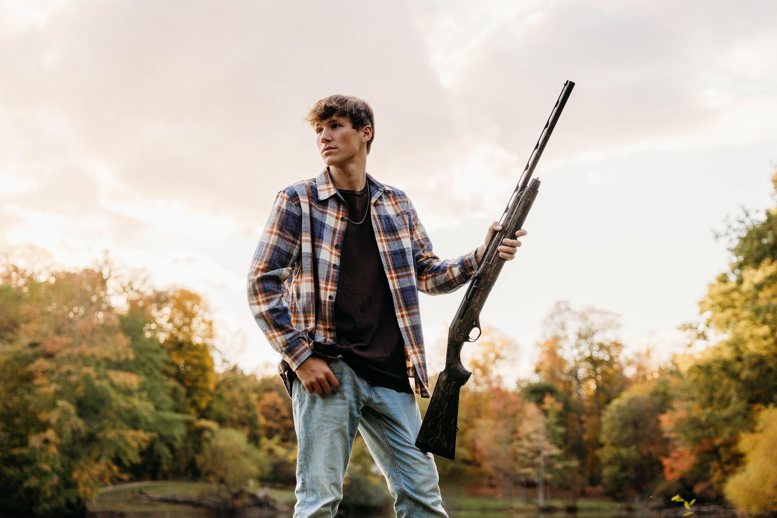 Hunter in plaid shirt and jeans holds rifle while walking through autumn field at sunset.