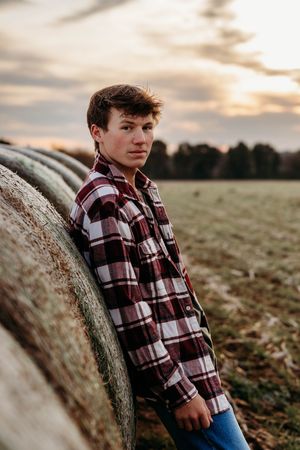 A person in a plaid flannel shirt leans against a hay bale in a field during golden hour sunset.