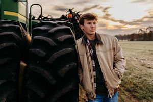 A person in a beige jacket stands in a field at sunset with dramatic clouds in the background.
