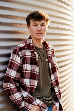 A young person in a red and white plaid flannel shirt and olive t-shirt poses against a corrugated metal wall.