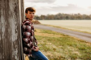 A person in a plaid shirt sits on a wooden fence post in a misty rural landscape at sunset.