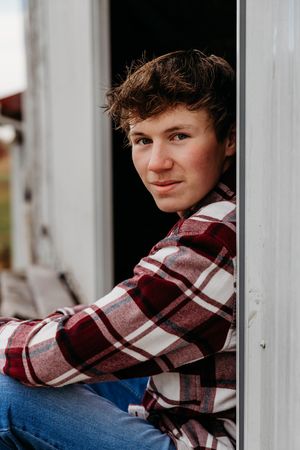 A person wearing a red and white plaid flannel shirt leans against white porch columns outdoors.
