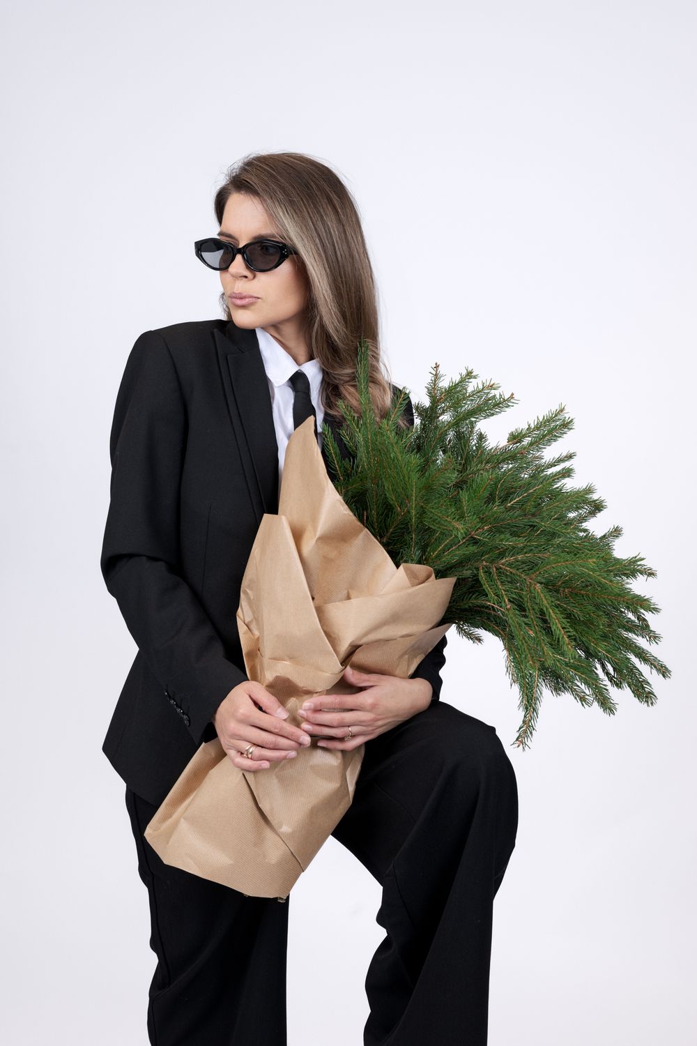 Person in black suit holding evergreen branches wrapped in brown paper against white background.