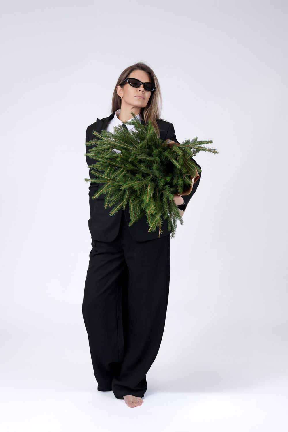 Figure in black suit and sunglasses poses with wrapped pine branches on white backdrop.