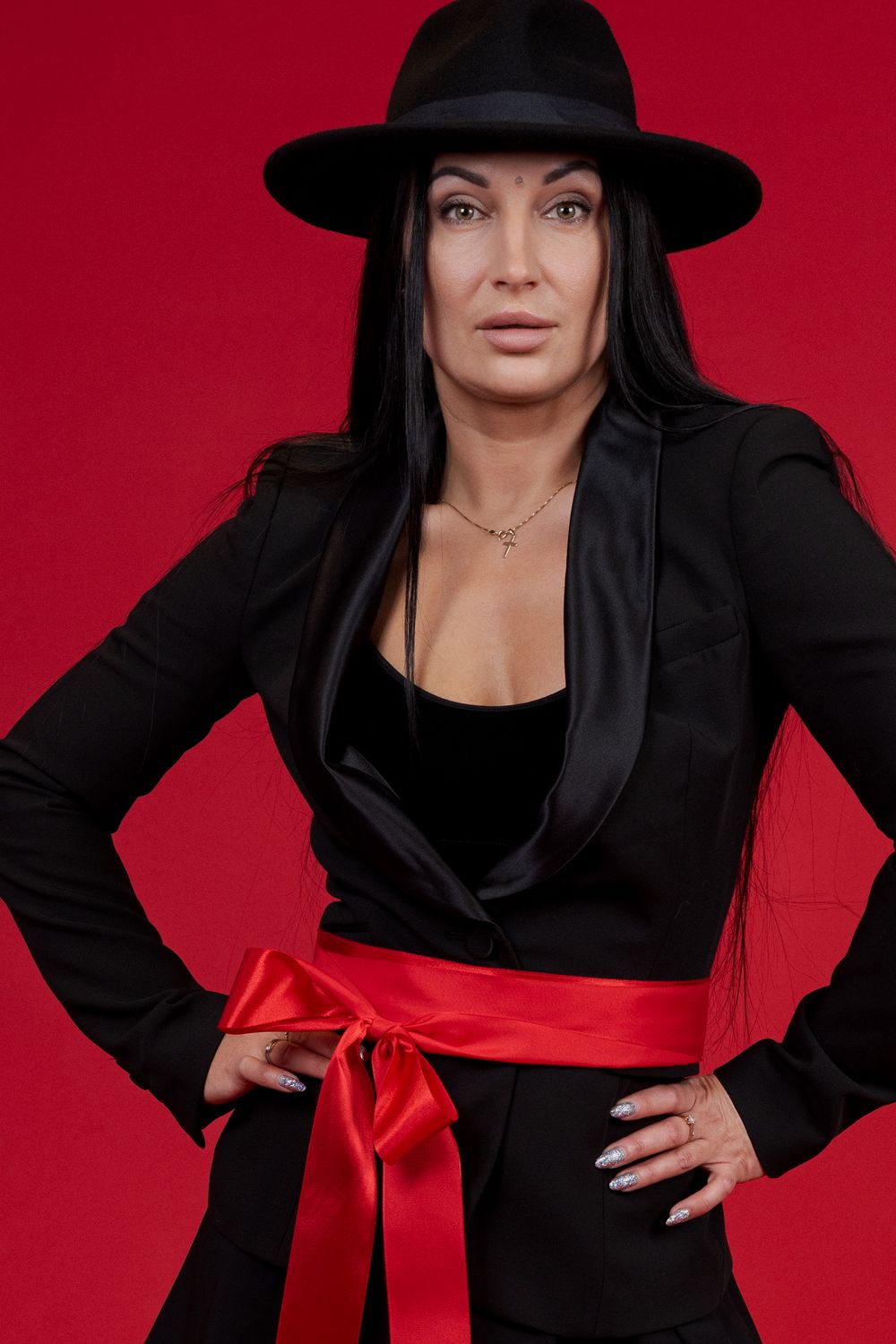 Figure in black outfit with wide-brimmed hat poses against red background with red ribbon accent.