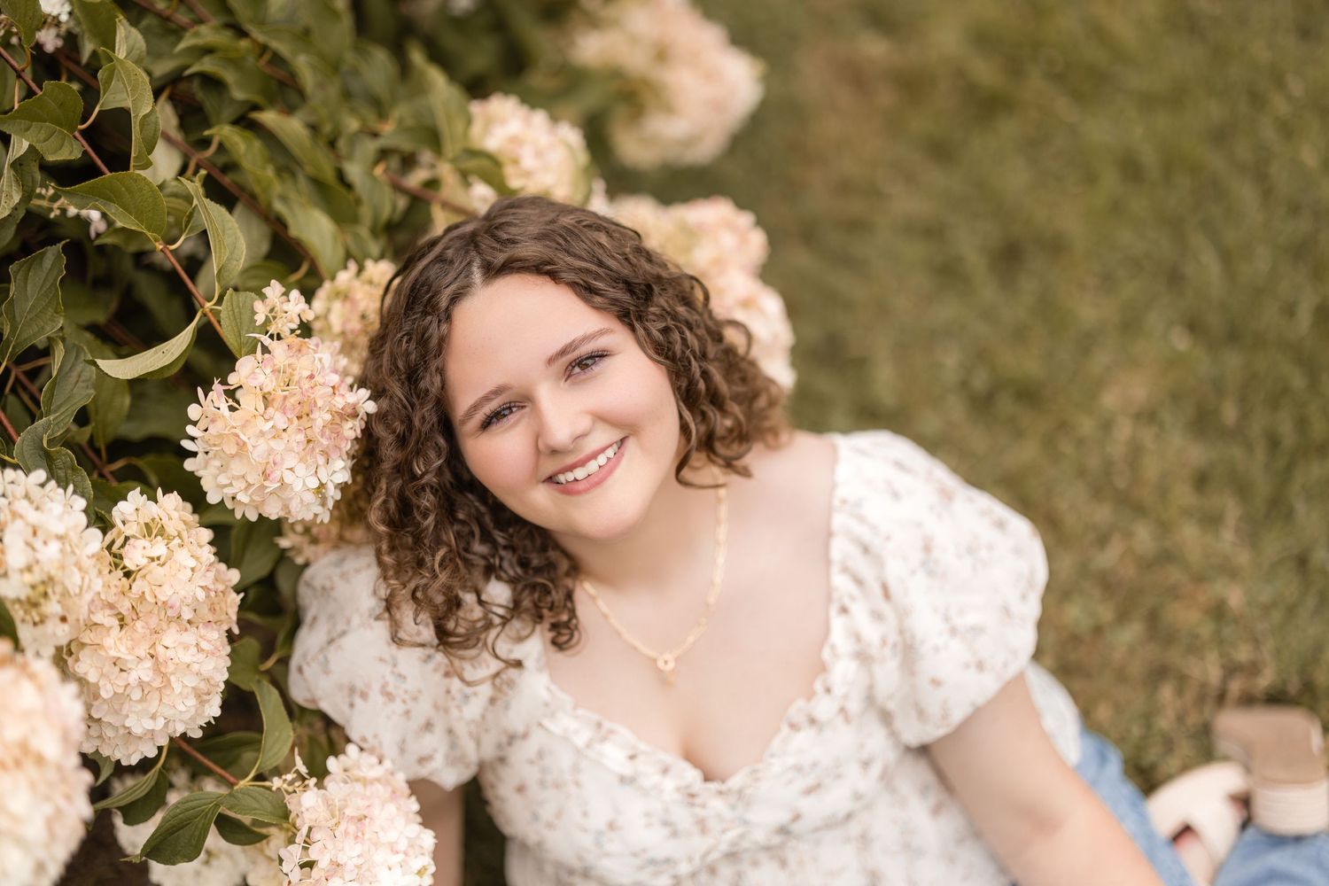 A person in a white lace dress poses near blooming peach hydrangea flowers in a garden setting.