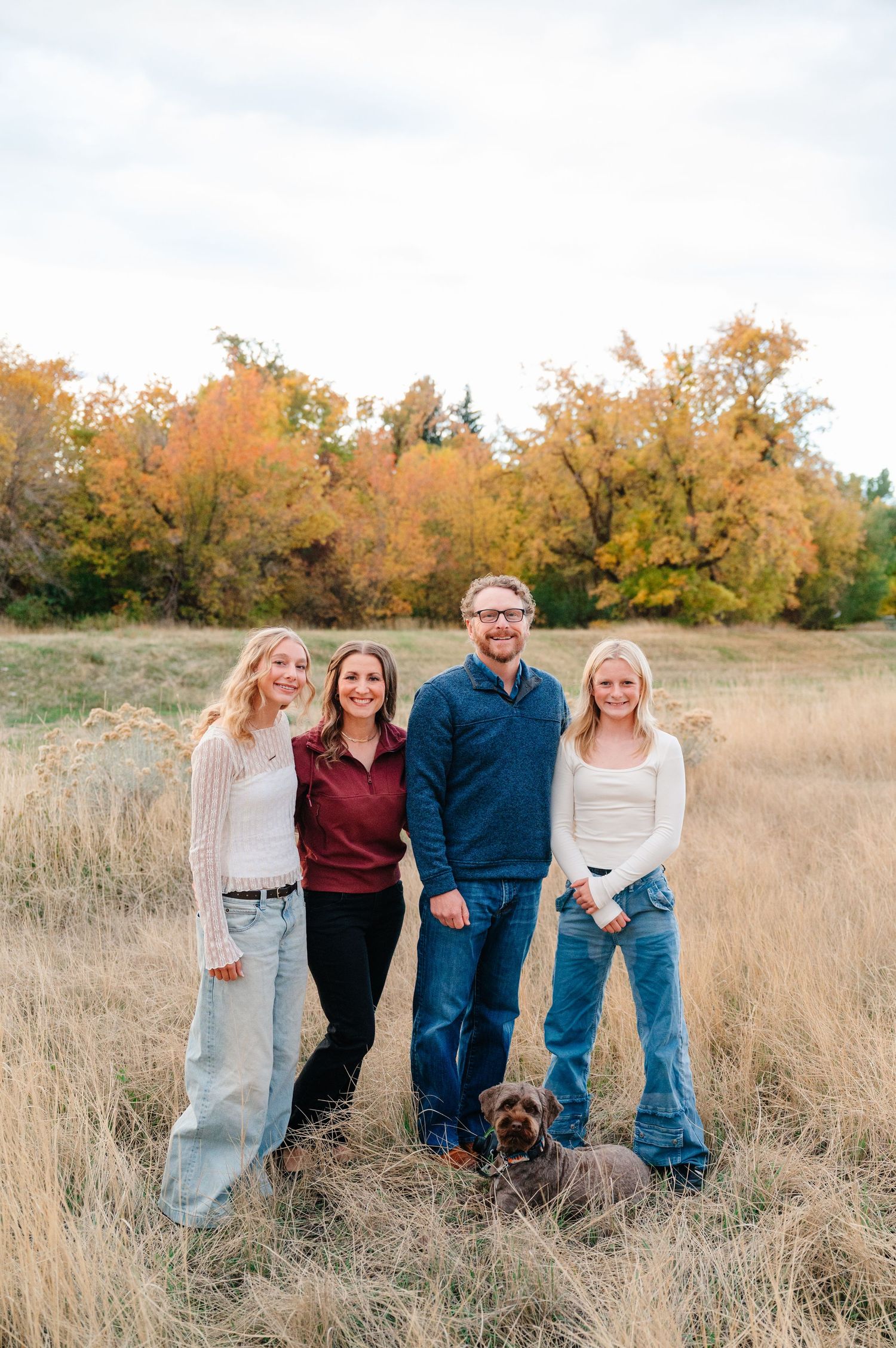 Family and their chocolate lab pose together in a golden autumn field during sunset.