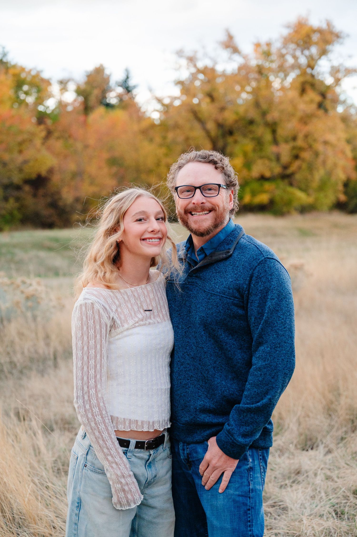 A couple in casual outfits smiling together in a golden autumn field during sunset.