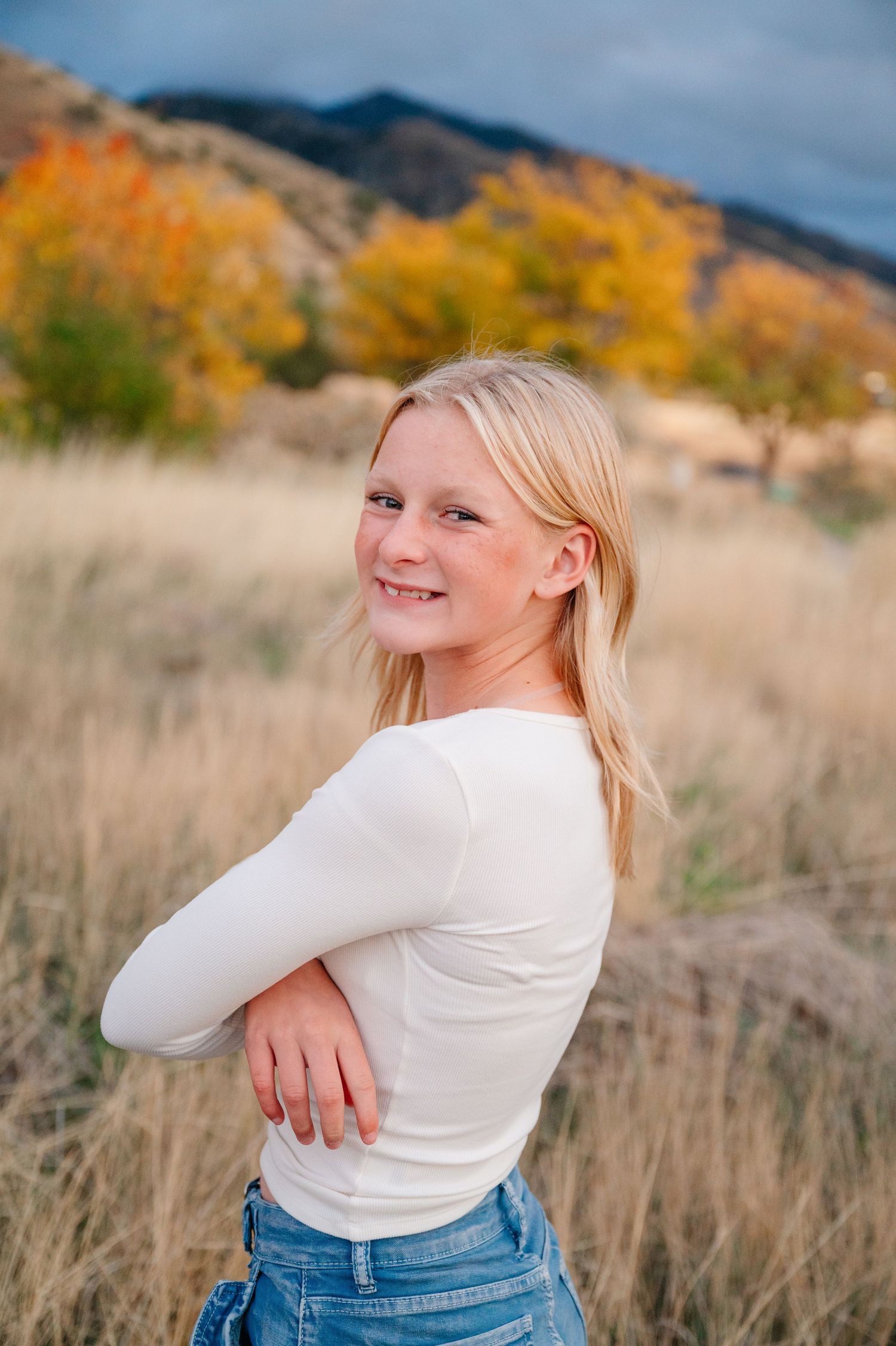 Person in white sweater and jeans poses in autumn field with mountains and fall foliage in background.