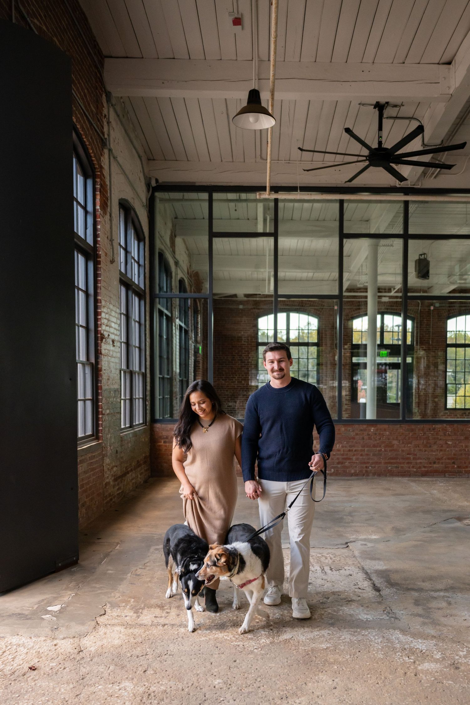 A couple and their dog stand in a spacious industrial room with exposed brick walls and large windows.