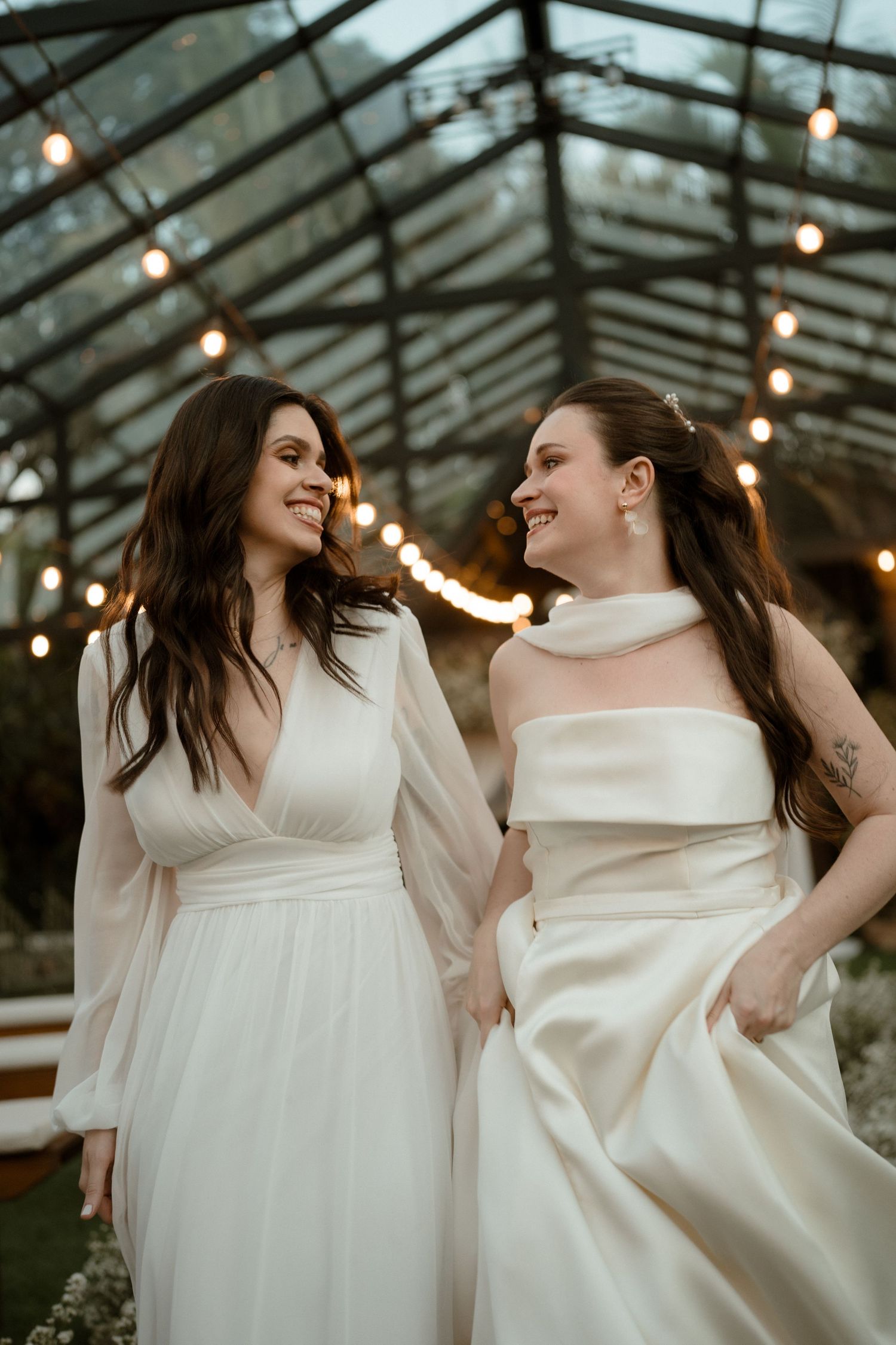 Two people in elegant white wedding dresses share a joyful moment under a greenhouse ceiling with string lights.