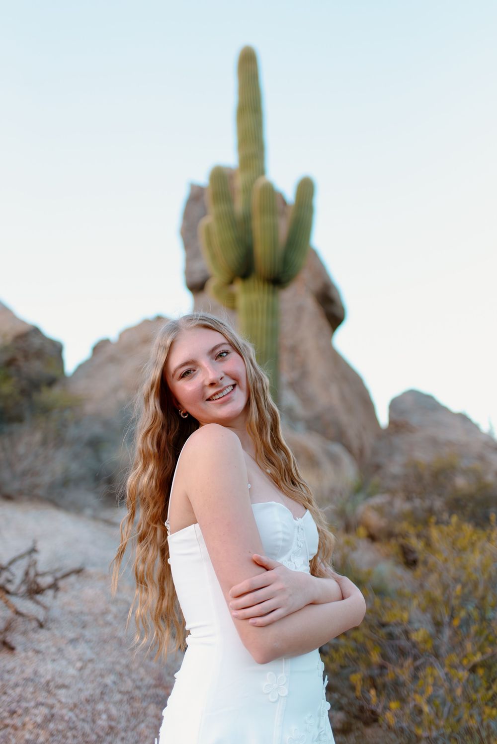 A high school senior in a white dress poses in front of desert rock formations and a tall saguaro cactus at sunset in Scottsdale, Arizona.