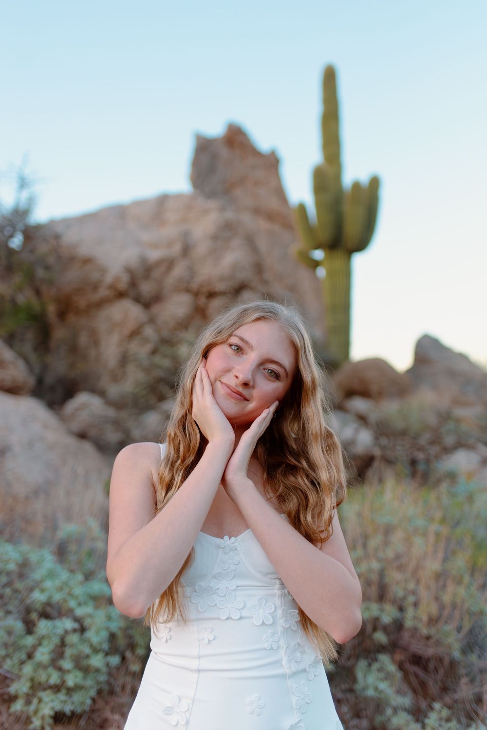 A high school senior in a white dress poses in front of desert rock formations and a tall saguaro cactus at sunset in Scottsdale, Arizona.