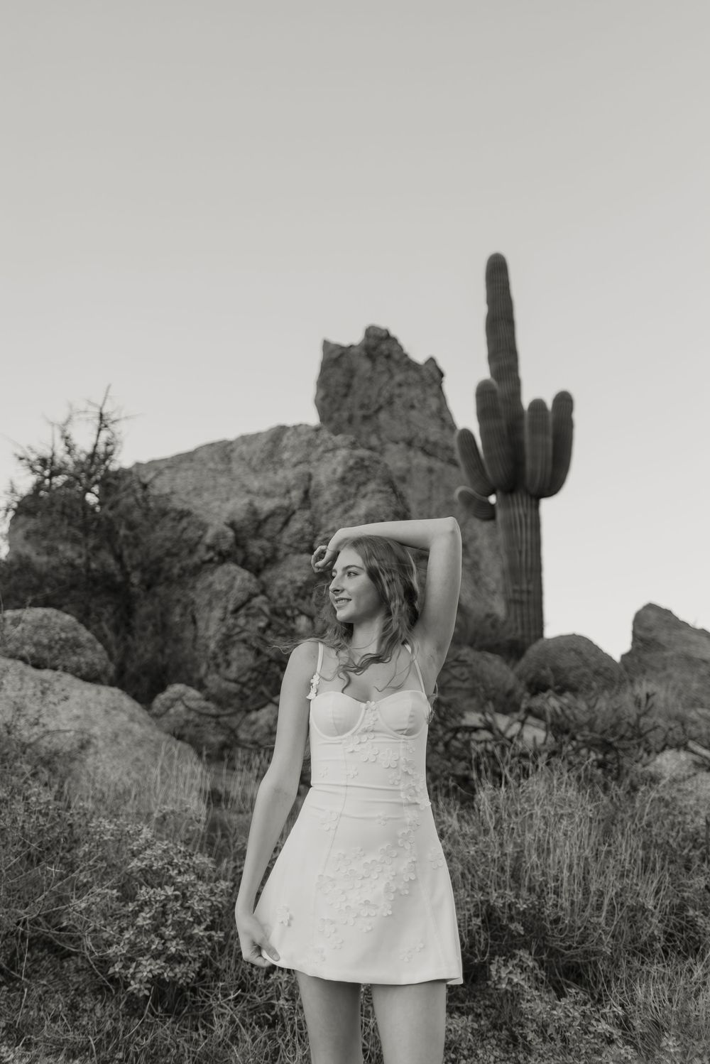 A high school senior in a white dress poses in front of desert rock formations and a tall saguaro cactus at sunset in Scottsdale, Arizona.