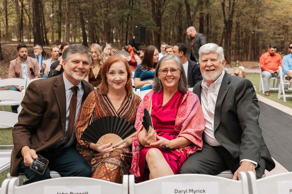Wedding guests sit together in white chairs at an outdoor autumn ceremony in a wooded setting.
