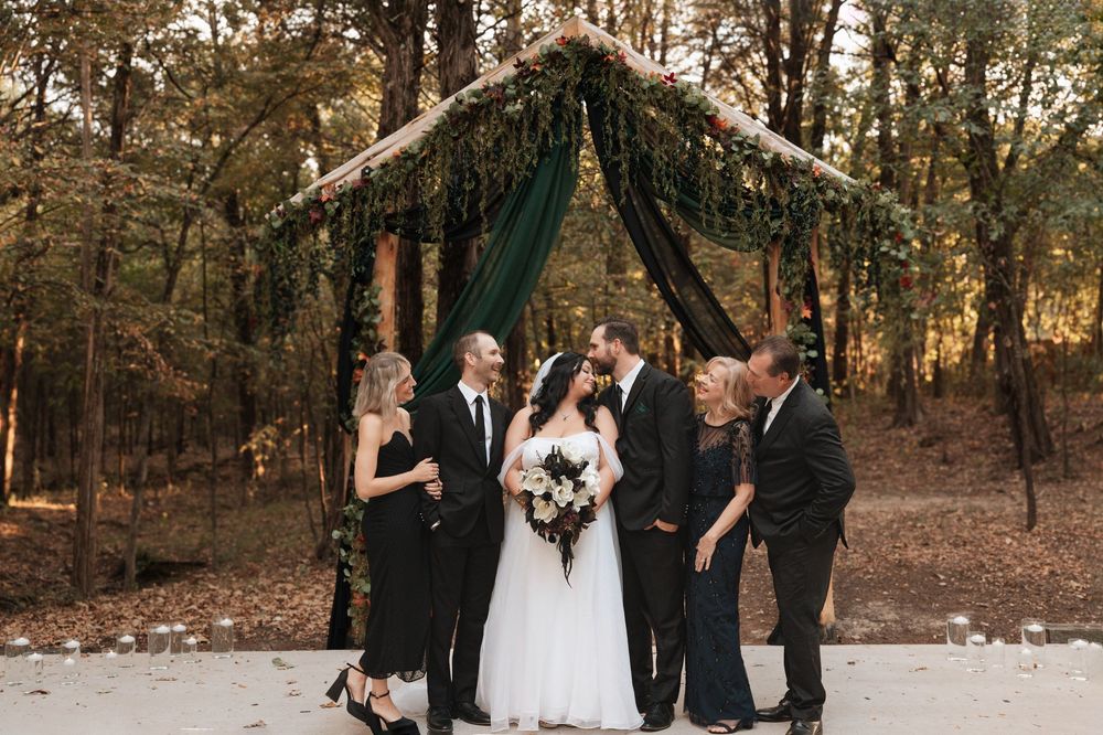 A wedding ceremony under a draped dark green canopy decorated with greenery in an autumn forest setting.